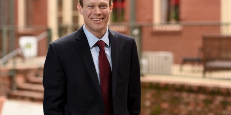 Adam J. MacLeod poses in a black suit and red tie.